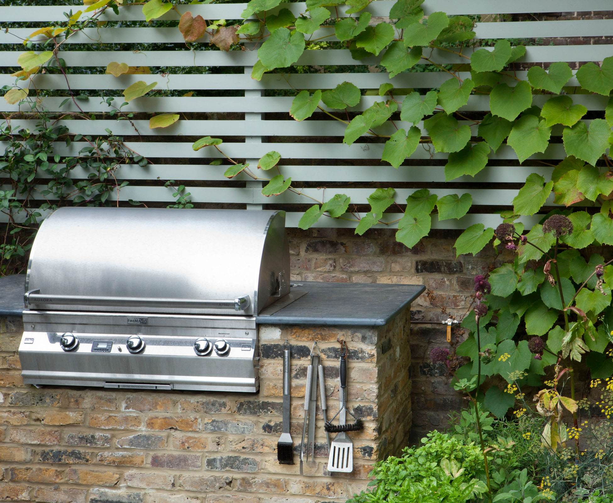 A stainless steel built-in barbecue has been installed by the landscapers in a brick wall, under a slatted trellis, painted light grey. A climbing vine is covering it.