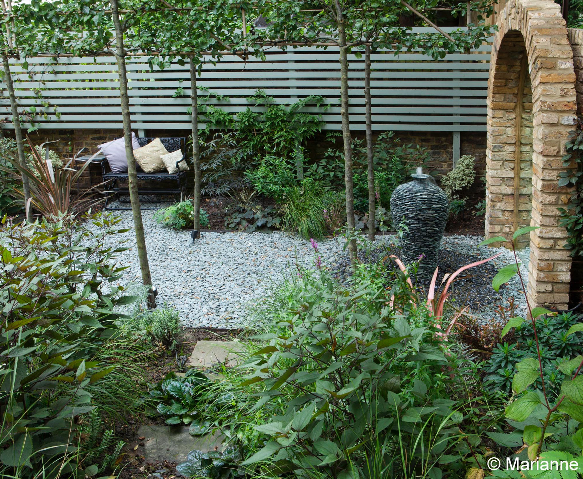 An avenue of pleached trees frames the gravel path to the mirror, where a slate water jar has been transformed into a bespoke water feature. 