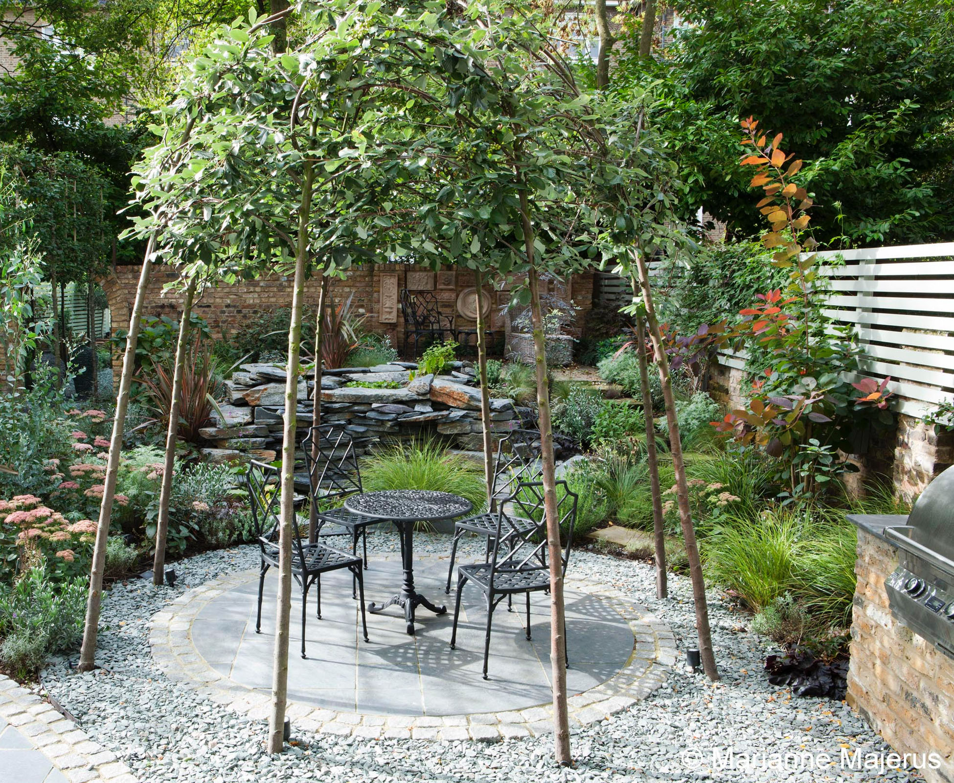 Some shade is provided by the trained deciduous trees above the slate patio, forming a cosy dinning area. A water feature in the back emits a nice constant sound of falling water in the distance in this Belsize Park garden.