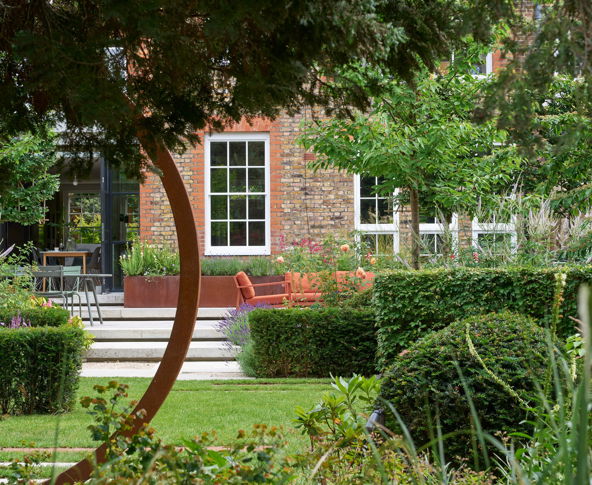 A long lens view through from the woodland to the house facade and patio, with staggered hedging and topiary for seasonal interest.