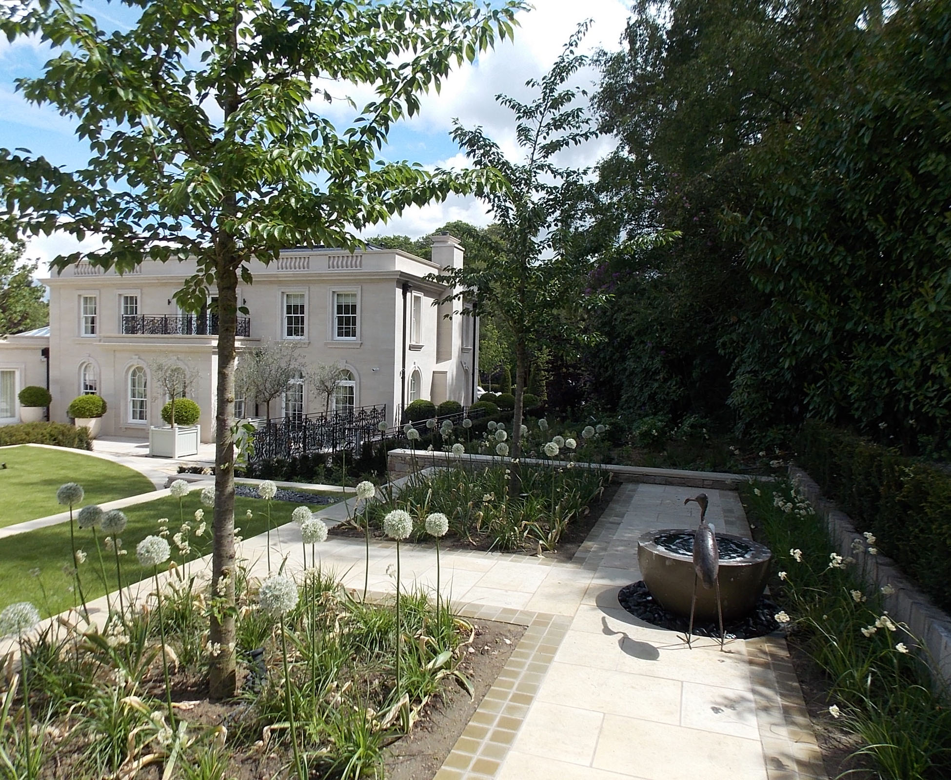 View towards this country house and garden, from the sandstone paving, where a water feature bowl is installed next to an iron sculpture bird. The cherry trees are underplanted with some white Allium.