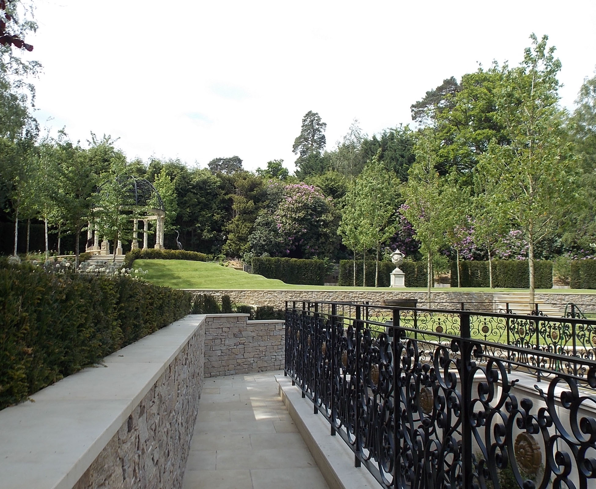 A view from the side passage, near the detailed gates, shows the retaining walls and sandstone paving leading to the garden. In the background, a lush and dense forest encloses the garden, offering good privacy in this country garden.