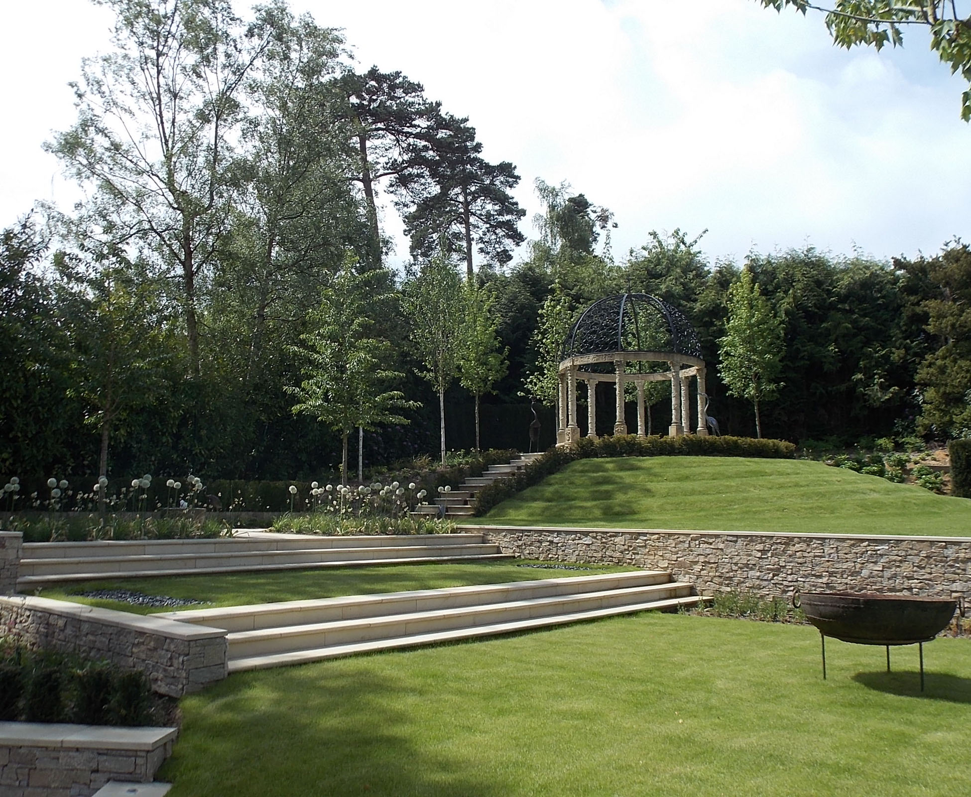 The designed pavilion is standing on the top of the hill, surrounded by low box hedges. A water feature, cascade, runs along the steps to a feature bowl, creating a lovely constant sound. Lower, on the lawn, stands an iron firebowl in this country garden.