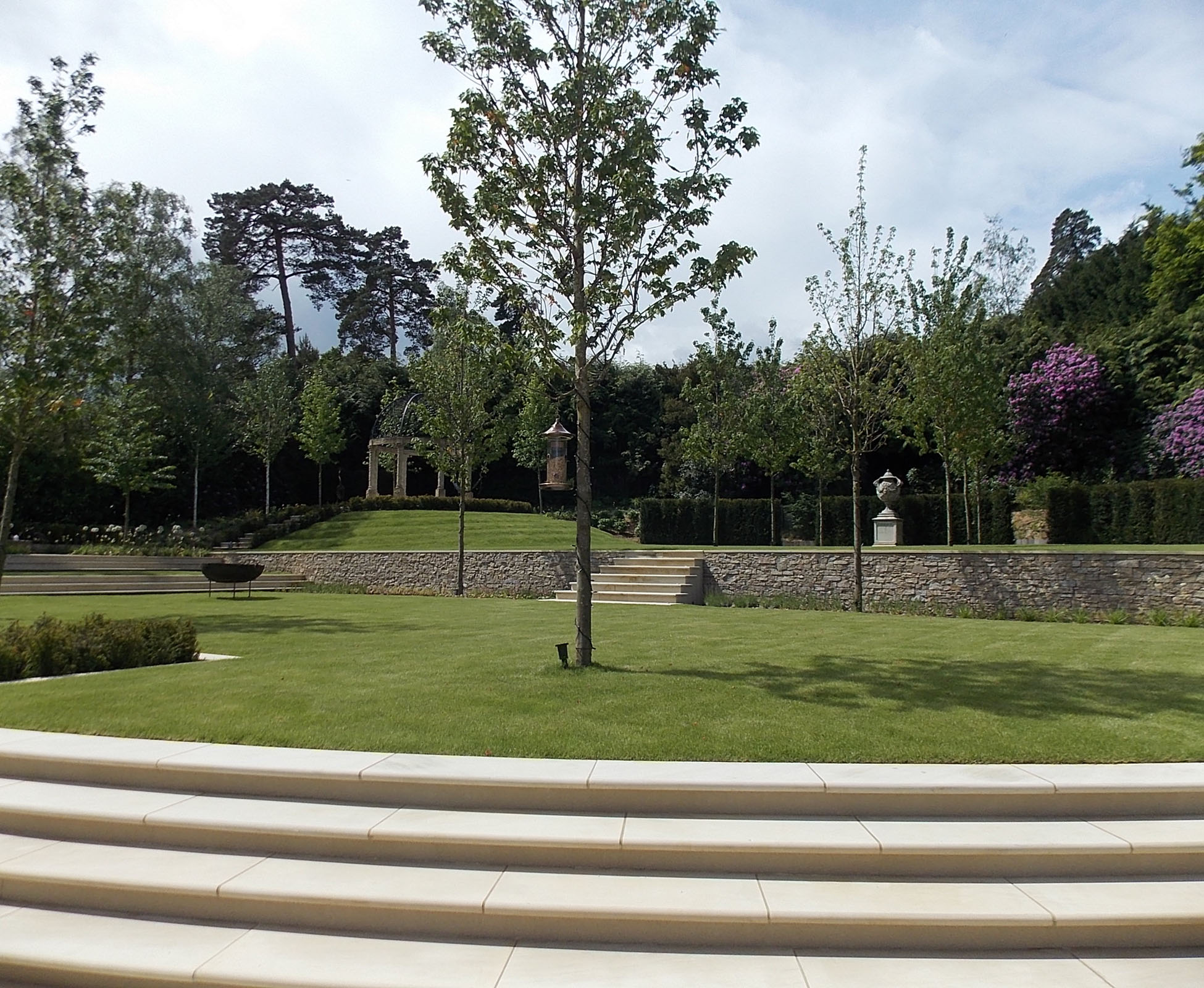 In spring, this country garden looks lush, with a big lawn planted with healthy maple trees. Some sandstone steps give access to the upper garden, retained by stone clad walls. At the back, a tall yew hedge provides separation from the forest entering the space.