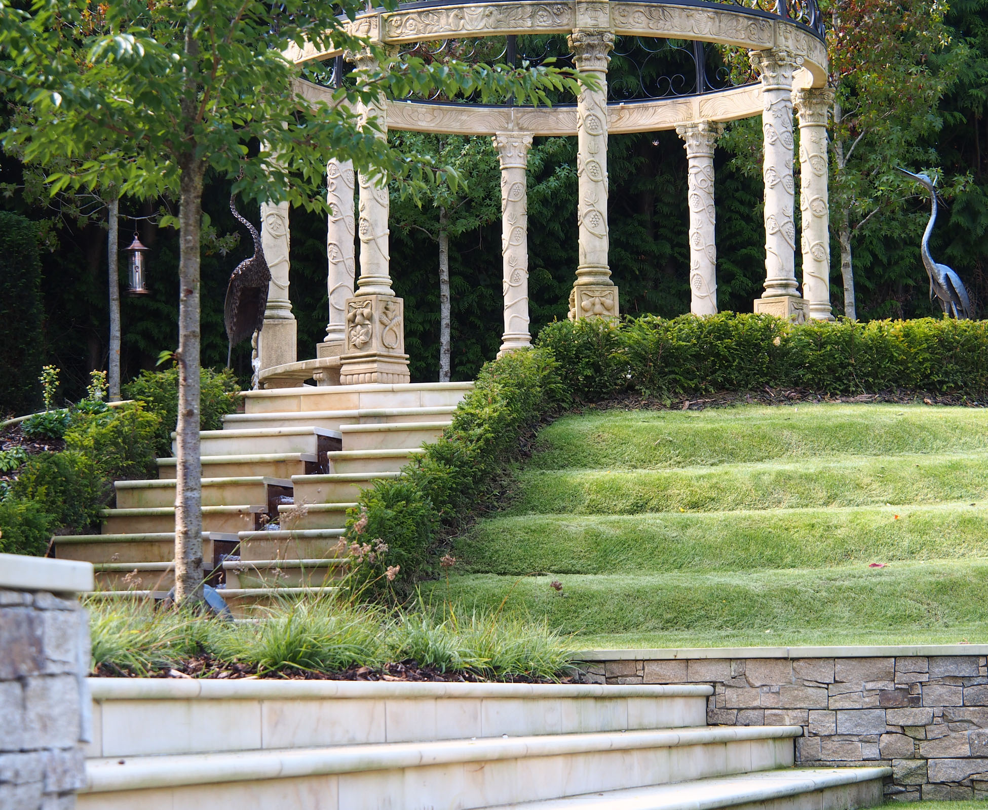 Close up of the bespoke pavilion, showing the arches and the steps down, with the water cascading to a feature bowl. An iron heron is standing in the lawn, behind a low evergreen hedge in this country garden.