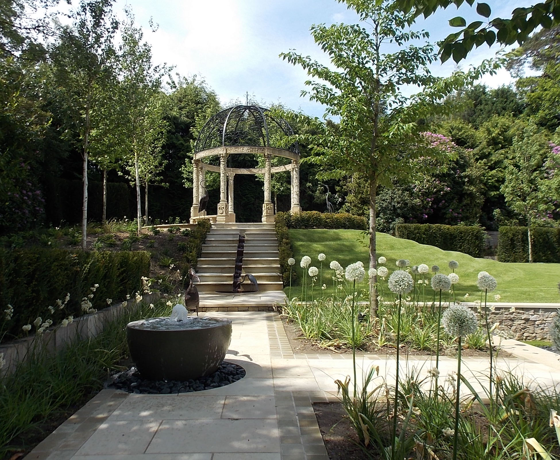 The pavilion has been built on the top of a hill, overlooking the cascade and water feature stepping down along a low evergreen hedge. Some cherry trees frame the view, underplanted with some white alliums in this countryside garden and house.