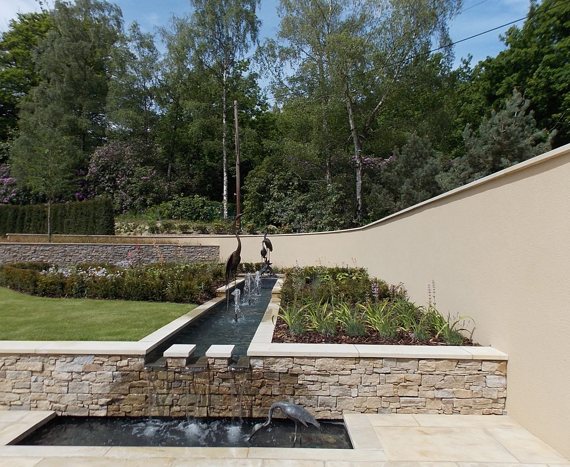 A second water feature is situated on the corner of the sandstone terrace, in full sun. The water cascades down to a basin, where some sculptures are installed. The beige rendered wall is softened by some herbaceous planting and grasses such as Miscanthus sinensis.