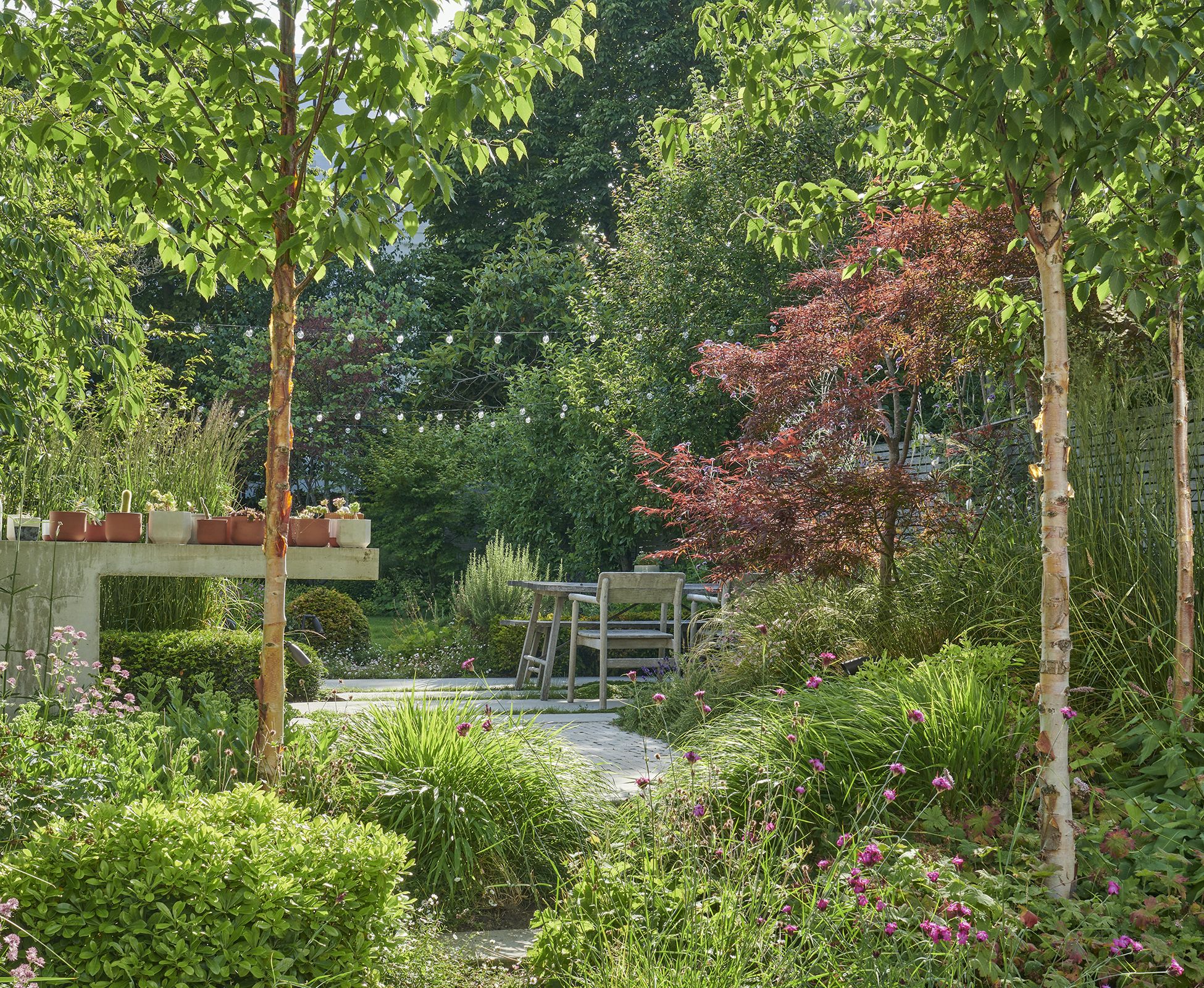A gorgeous view of the rear garden, featuring a bespoke concrete kitchen counter, dining area, and a path leading down the lawn, all surrounded by lush planting.