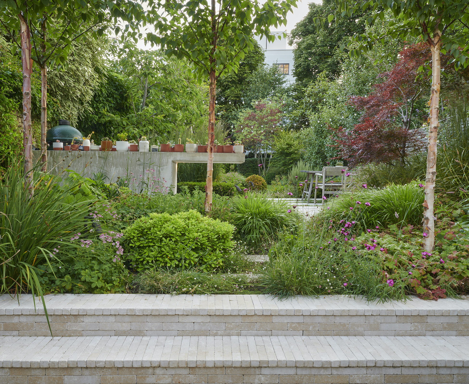 Two steps clad in stone pavers lead from the patio up to the main garden level with lush plants around them softening the transition.