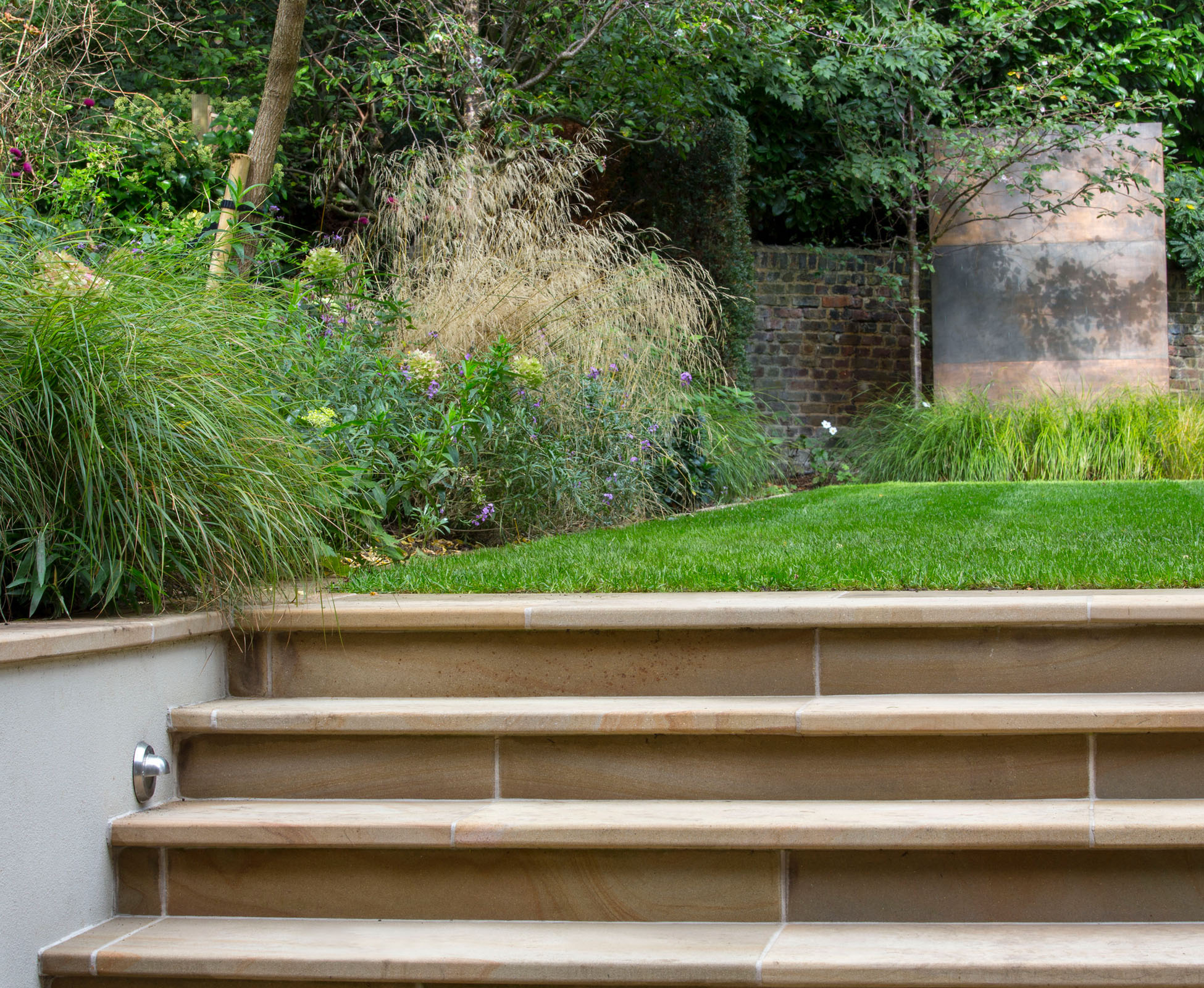 A buff rendered wall, with yorkstone coping detail, is planted with overhanging herbaceous perennials and grasses to soften the edges of this Hampstead Heath garden.