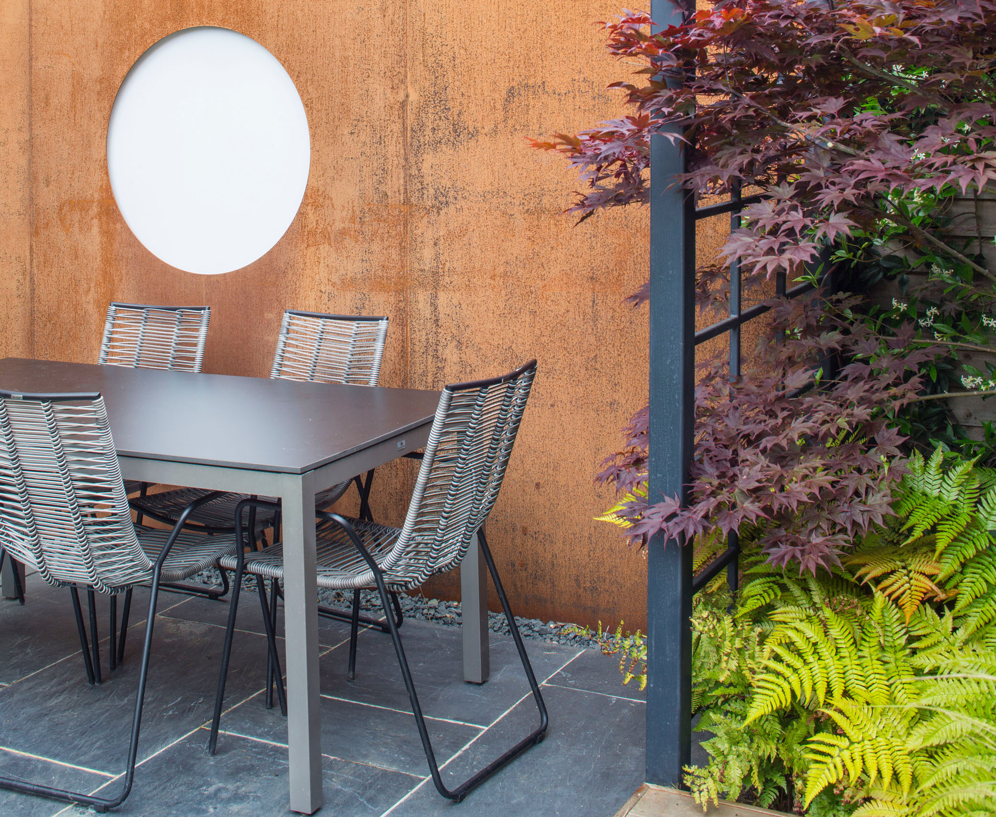 A family size table and chairs are installed on the blue slate paving, near the Corten steel panel feature. A powder coated frame creates a translucent division, with the purple foliage of Acer palmatum ‘Atropurpureum’ coming through.