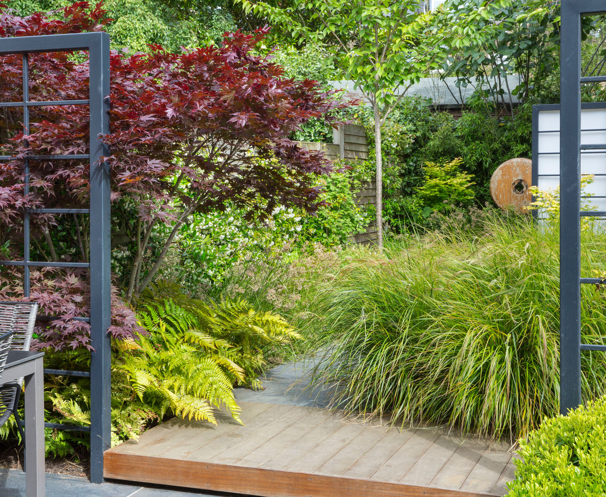 The lush planting, mainly evergreen, overhangs the hardwood deck, accentuating the main path to the back of this Queen’s Park garden. The eye is attracted to the wooden sculpture sitting in front of a tall bamboo screen.