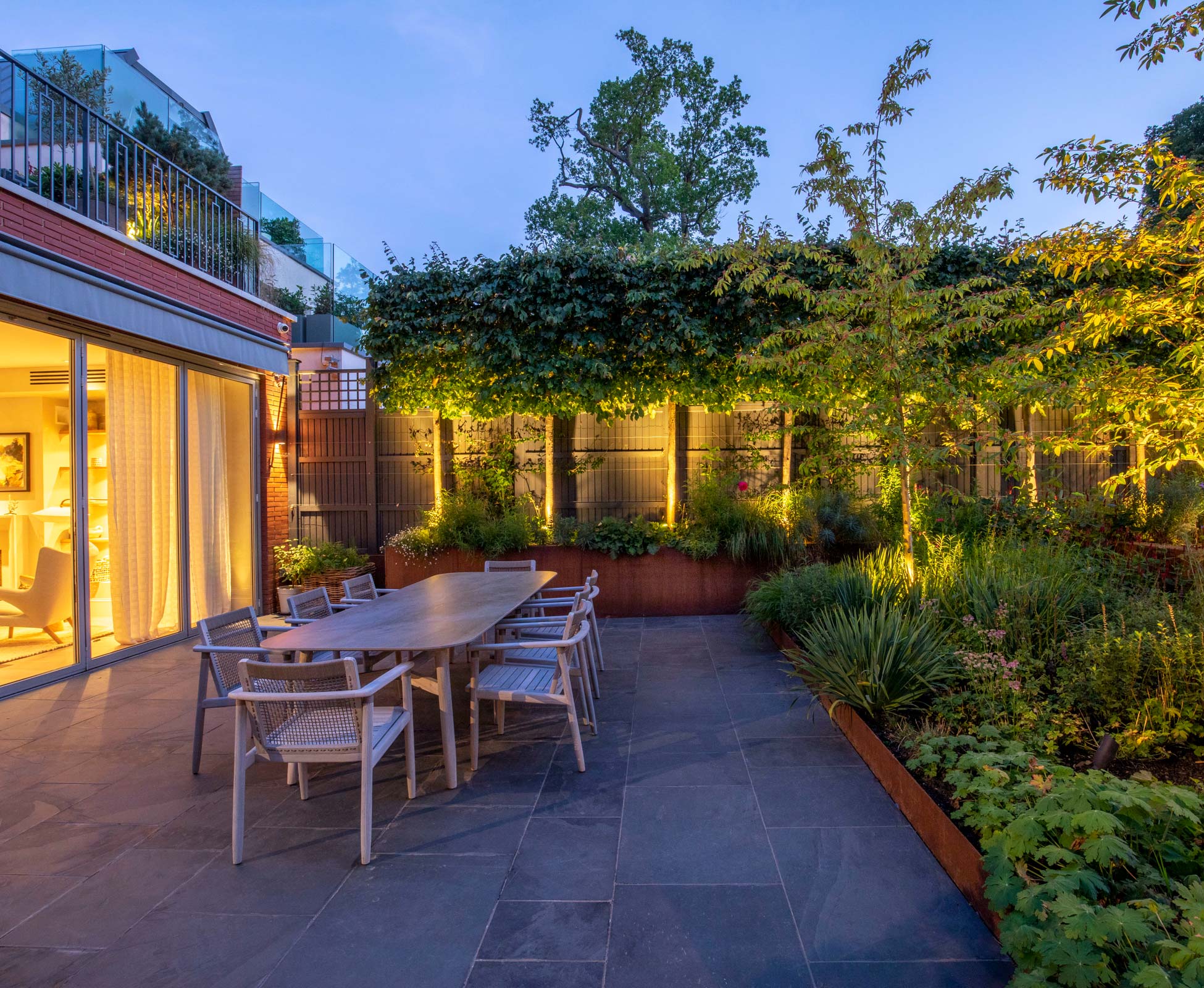 At dusk pleached Parrotia and Cotoneaster cornubia trees are lit up with a warm white LED to create a subtle and elegant atmosphere around the dining patio area.